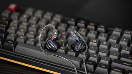 Nærbillede i trekvart fugleperspektiv af et par sorte, matmetal in-ear-høretelefoner med iriserende slidser på forsiden, flettet grå metalledning og gennemsigtige stik med rød og blå ring, placeret på et gråt mekanisk tastatur