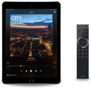 Tablet, der viser et billede af en oplyst by om natten, og sort Naim Audio-fjernbetjening med drejeknap og knapper