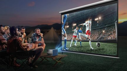 6 cilvēku grupa sēž uz saliekamajiem krēsliem, projekcijas ekrāns rāda futbola spēli, projektors uz zema galdiņa