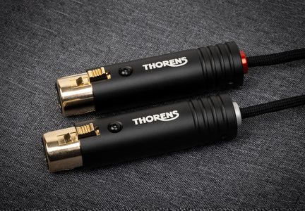 Dva kovové matně černé XLR konektory typu samec s bílým logem THORENS, pozlacenými špičkami a identifikačními kroužky v červené nebo stříbrné barvě, pohled shora na textilním šedém pozadí