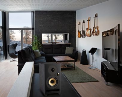 Modernus salonas su tamsiu kampiniu sofa, mediniu kavos staliuku, ant sienos kabančia gitara, dideliu langu, juoda kolonėle pirmame plane.