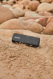 Trådløs Sonos Roam Shadow Black Bluetooth-højttaler placeret på sand, omgivet af småsten