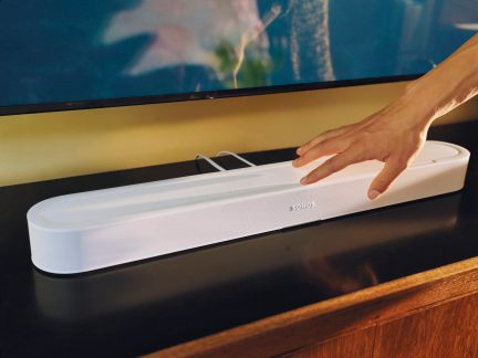 Hvid Sonos-soundbar placeret under et fjernsyn med en hånd, der betjener de øverste berøringsknapper