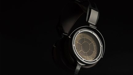 Svart, rundt-øret-hodesett i metall og lær, åpne ørekopper med gyllen grill og synlige skruer, sidevisning, synlig kabel