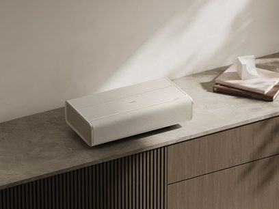 XGIMI Aura 2 stačiakampis vaizdo projektorius, šviesi apdaila, pastatytas ant medinio baldo, sukrautos knygos, geometrinė skulptūra