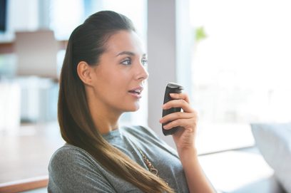 Ung brunette kvinde set fra siden, der holder en sort smartphone tæt på sit ansigt og kigger på skærmen i et lyst interiør