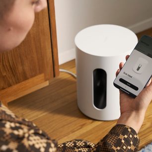 Mat beyaz Sonos Sub Mini bas hoparlörü, bir elde tutulmuş şekilde, önden görünümle ahşap bir mobilyanın önünde