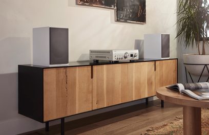 Denon PMA-1700NE sølvfarget hi-fi stereoanlegg, frontvisning, på trehylle, med grå høyttalere og dekorative gjenstander