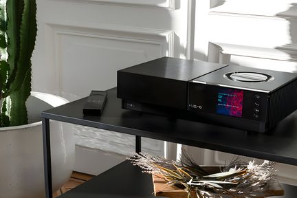 Melns audio pastiprinātājs ar zilu LED displeju uz koka galda, blakus vāzei ar sausām zarām