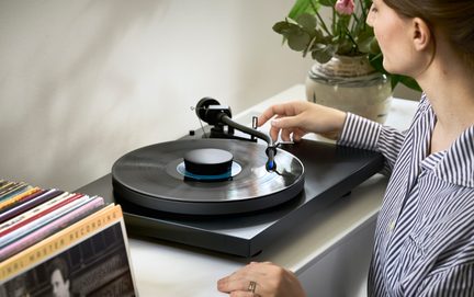 Černý gramofon s vinylovou deskou, ruka nastavující rameno, pruhovaná košile, váza s květinami, řada desek vlevo