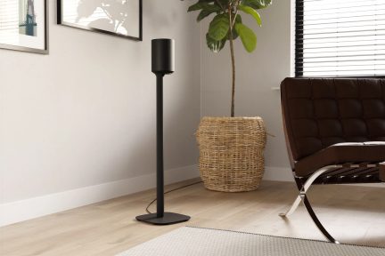 Black speaker on a slim stand with a flat base, in a living room next to a brown armchair and a large green plant