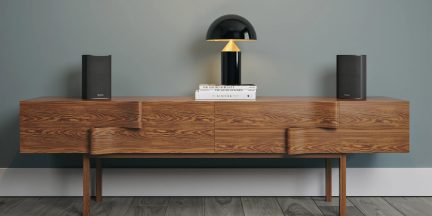 Buffet in legno con 2 altoparlanti neri, 3 libri impilati e lampada nera e gialla su sfondo di muro blu