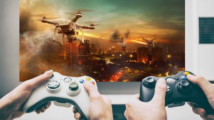 To hænder, der holder gamingkontroller foran en by i flammer med droner, der flyver i en skumringsfarvet himmel