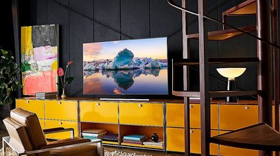 Samsung QE75Q85R-TV som viser et bilde av isfjell speilet i vannet, plassert på et gult møbel i et rom