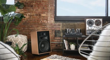 Højttaler i brunt træ med stor woofer og horn, sort front, set fra tre kvart, op ad en murstensvæg