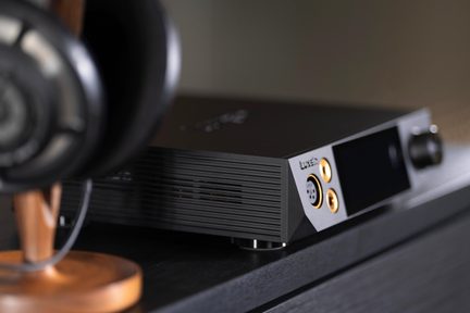 Fekete hi-fi fejhallgató-erősítő aranyozott jack csatlakozókkal a bútoron, elöl életlen fa tartón lévő fejhallgatóval