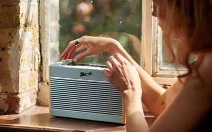 Retro přenosné rádio v pastelově modré barvě z žebrovaného plastu, pohled ze tří čtvrtin zepředu, umístěné na parapetu, ruka otáčí horním knoflíkem