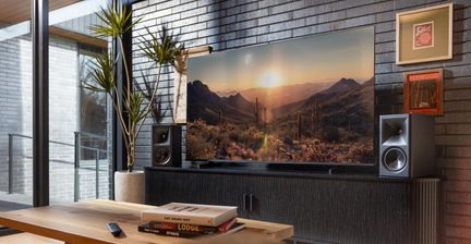 Großformatiger Flachbildfernseher, der eine Wüste bei Sonnenuntergang zeigt, zwischen zwei Lautsprechern auf einem schwarzen Möbelstück