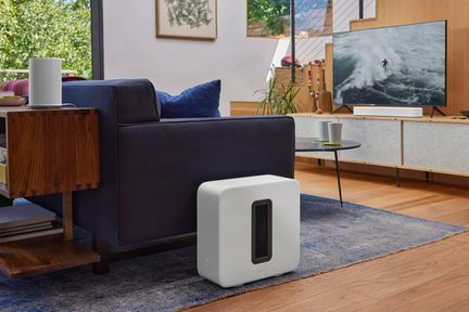 Hvid Sonos Sub subwoofer placeret på gulvet ved siden af en blå sofa i en moderne stue med tv og soundbar