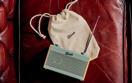 Bärbar rektangulär radio i pastellgrönt plast med rillad frontgrill, utdragen teleskopantenn i metall och beige sidor, uppifrån vy, placerad på rött läder bredvid en ekofärgad tygpåse med snöre