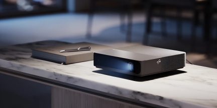 Videoproiettore a ottica ultra corta nero e grigio visto di tre quarti su tavolo in marmo con fascio luminoso