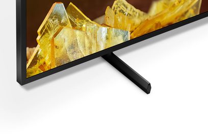 Sony XR98X90L TV, set fra en vinkel, sort ramme, skærm viser gule krystaller, sort metalfod, hvid overflade