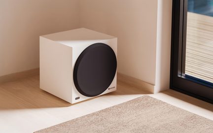 Valkoinen, mattapintainen, maalatusta puusta valmistettu kuutionmuotoinen subwoofer, jonka etupuolella on suuri musta pyöreä kaiutin ja alareunassa pieni suorakulmainen LED-valo, kuvattuna etuviistosta