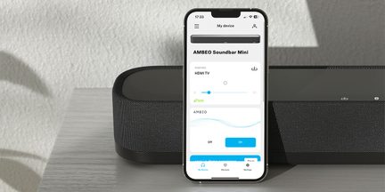 En smartphone, der viser en app, er placeret foran en sort soundbar på en lys overflade med en tekstureret væg i baggrunden
