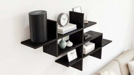 Designová tmavě černá nástěnná police s posunutými přihrádkami, na níž jsou knihy, hodiny, vázy a černý reproduktor.