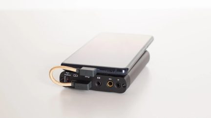 Bærbar grå metal-forstærker-DAC, 3,5- og 4,4 mm-stik, flettet USB-kabel, smartphone placeret ovenpå, trekvart vue