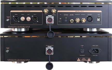 Vista trasera, dos cajas de audio apiladas en metal negro mate con ribetes dorados y pies cilíndricos plateados, que presentan múltiples conectores XLR, RCA, BNC, ópticos y USB, así como tomas de alimentación circulares protegidas por una tapa flexible