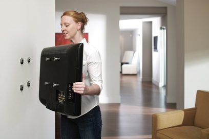 Kvinde set fra siden, der holder en sort vægbeslag til tv i et moderne indendørsmiljø med sofa og åben dør i baggrunden