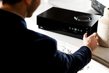 Vīrietis uzvalkā pielāgo melnu, taisnstūrveida elektronisko ierīci ar pogām, kas novietota uz baltas virsmas