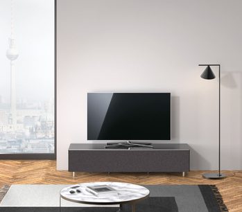 Lavt, mørkegråt TV-møbel med skuffe, på metalben, med fladskærm ovenpå, sort lampe ved siden af, set forfra