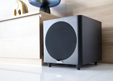 Küp şeklinde siyah Artsound subwoofer, önde büyük dairesel ızgaralı, açık renk ahşap dolabın önünde zemine yerleştirilmiş