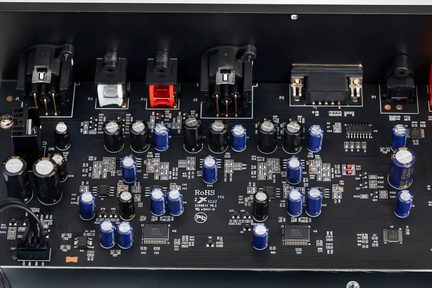 Sort epoxy-printkort set fra oven med mange blå og sorte cylindriske kondensatorer, flade SMD-chips og sorte plastikstik samt en rød og en hvid indsats