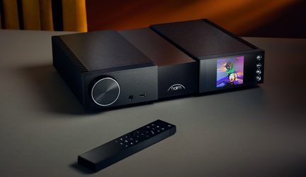 Czarny metalowy komponent hi-fi, widok 3/4, duży pokrętło, kolorowy wyświetlacz po prawej stronie, port USB i gniazdo słuchawkowe, pilot przed urządzeniem