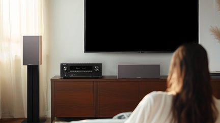 Svart veggmontert TV og møbel i brun tre, svart forsterker, grå senterhøyttaler i stoff og gulvhøyttaler, sett fra forsiden