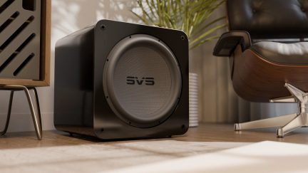 Subwoofer nero lucido con griglia circolare grigia e logo centrale, vista in prospettiva in un interno moderno