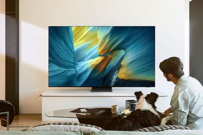 Televisore a grande schermo con cornice in metallo nero ultrasottile, pannello in vetro lucido, base centrale rettangolare, visto di fronte in un soggiorno luminoso