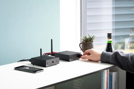 Twee zwarte rechthoekige kastjes met antennes, zwarte smartphone, hand die een knop aanpast, groene kamerplant in pot, donkere fles