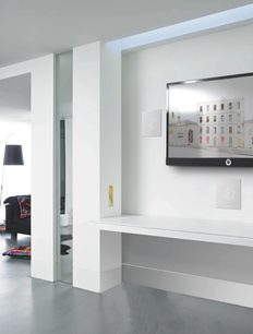 Moderní bílý interiér s nástěnnou televizí, barevným obrazem, prosklenými dveřmi a dekorativními lištami na stropě a stěně