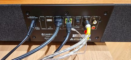 Bagside af en sort metal audioforstærker med lyst trætop, panel med tilslutninger til strøm, USB, HDMI ARC, RJ45, optisk og RCA, som allerede er forbundet med flere kabler