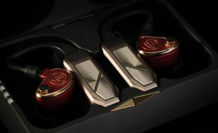 Oversigt over et sæt bestående af to røde in-ear-høretelefoner i metal med gyldne, prægede plader og knap, forbundet til to rektangulære champagnegrå moduler i børstet legering med skarpe kanter, placeret i en sort skumholder