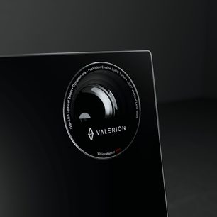 Zbliżenie pod lekkim kątem na czarną, błyszczącą, szklaną fasadę projektora Valerion VisionMaster Max, ukazujące wyraźny, okrągły obiektyw otoczony czarną, sitodrukowaną ramką