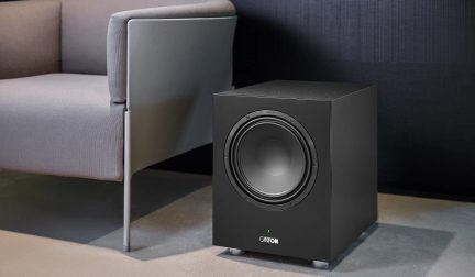 Zwarte Canton-subwoofer, driekwart van voren gezien op de vloer naast een fauteuil in een moderne woonkamer