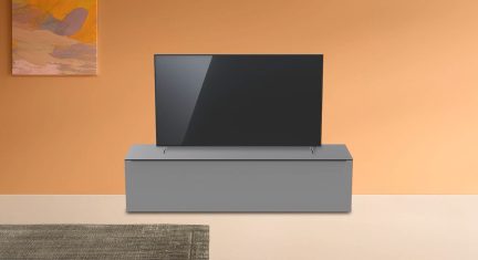 Televizor tmavě šedé barvy na světle šedém nábytku před oranžovou zdí s abstraktním oranžovým a šedým obrazem