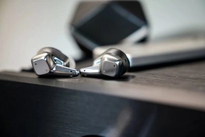 Vue en trois-quarts de deux écouteurs intra-auriculaires sans-fil Hifiman Svanar Wireless finition métal argent poli, coque anguleuse avec tige courte et évents discrets, posés sur une surface sombre