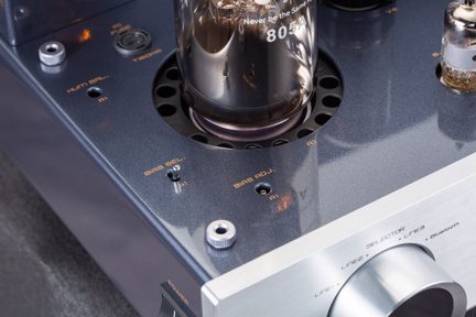 Tříčtvrtinový pohled shora na hi-fi lampový zesilovač s lesklým kovovým šasi v tmavě modré barvě a oranžovým potiskem, velkou skleněnou elektronkou označenou „805A“ zasazenou do kruhové perforované mřížky, doplněnou malými rýhovanými stříbrnými tlačítky, přepínačem biasu a hliníkovým ovladačem hlasitosti s kartáčovaným povrchem.