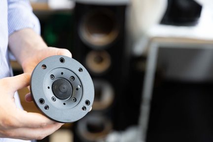 In de hand gehouden ronde zwarte hifi-luidspreker-tweeter, voorkant met schroefgaten, onscherpe luidsprekers op de achtergrond